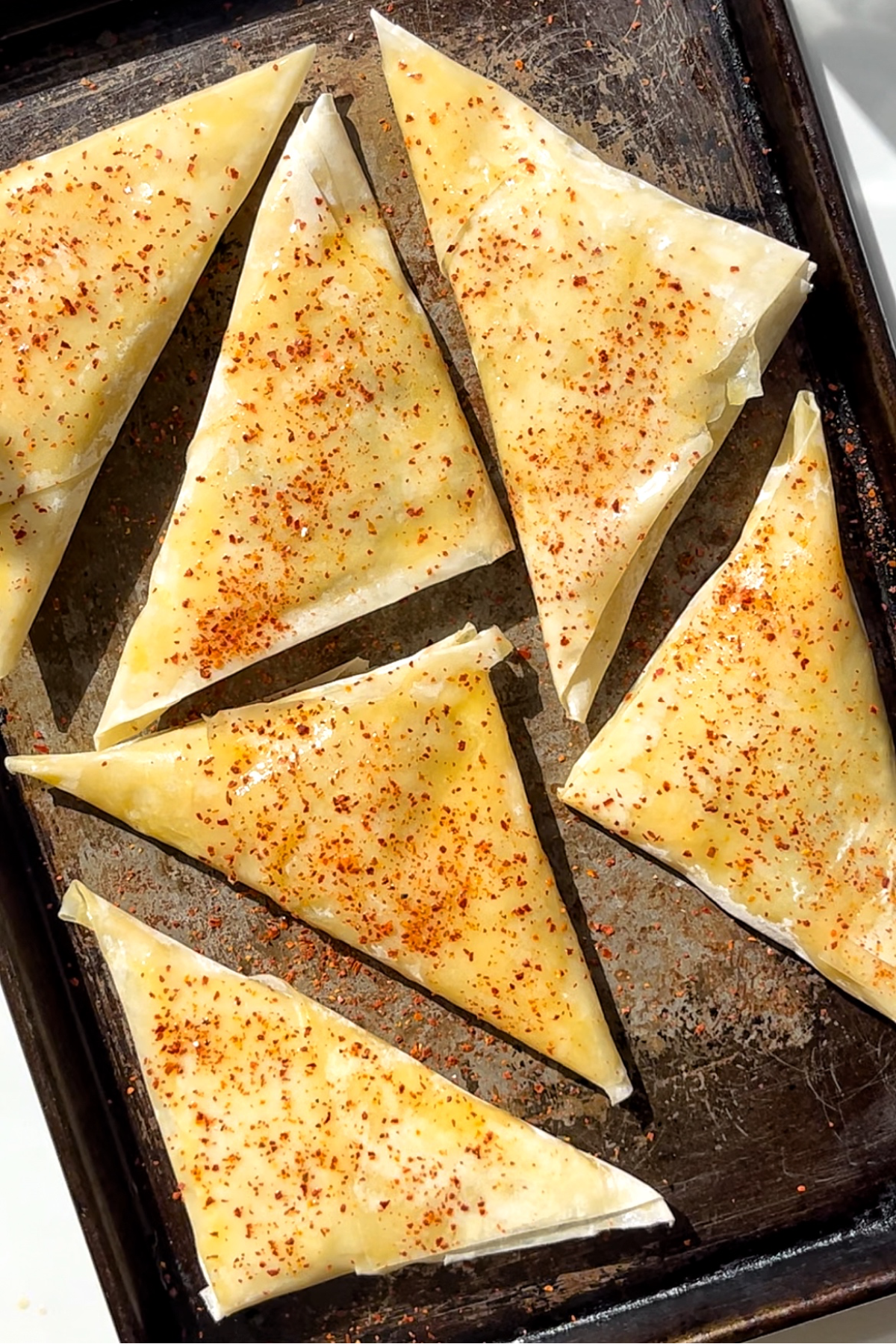Triangular pastries on a black baking tray sprinkled with chilli flakes.