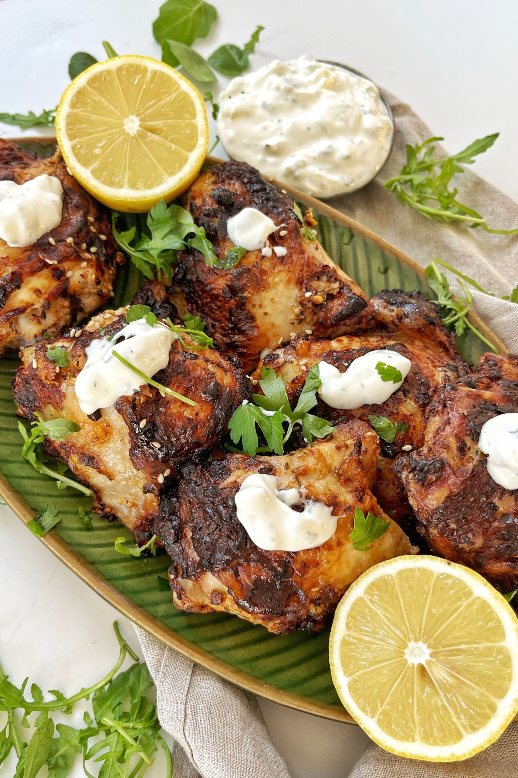 Cooked chicken thighs on a green plate with lemons and tzatziki.