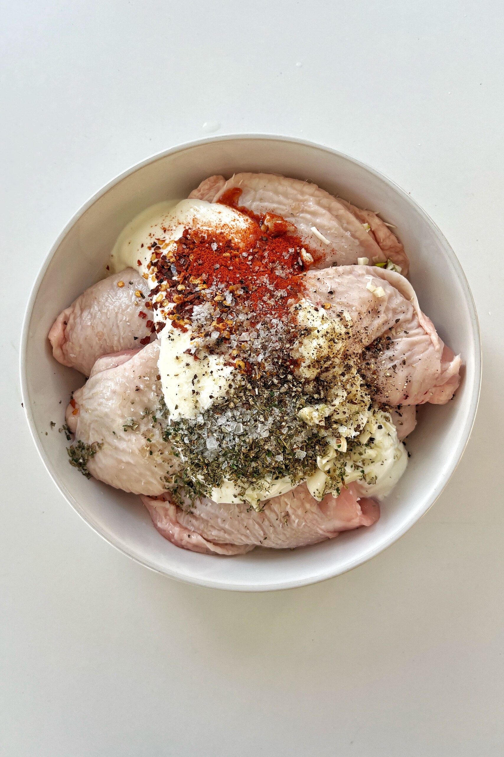 Bowl full of chicken thighs with yogurt, mixed herbs, paprika, and chilli flakes on top.