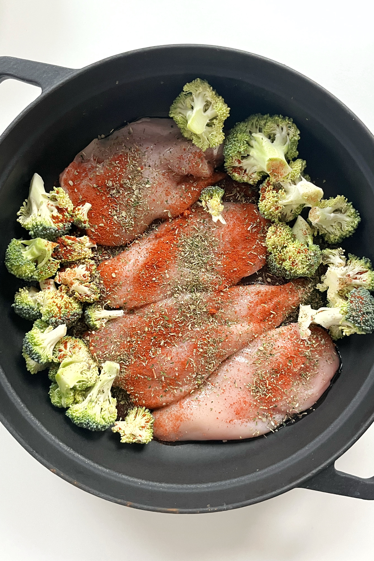Black pan with 4 pieces of chicken breast, broccoli, paprika, and mixed herbs drizzled over.