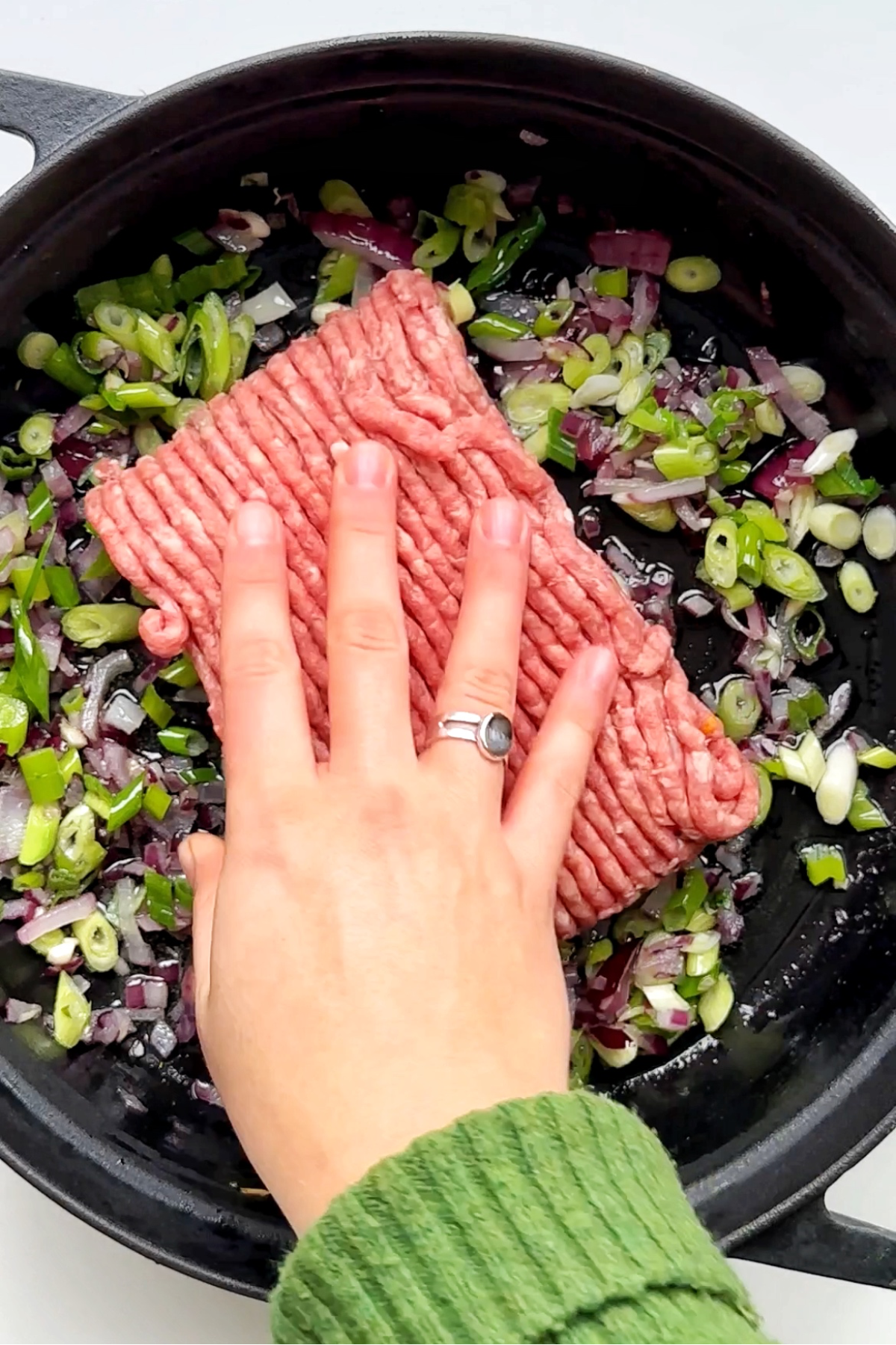 Black casserole pan with onions, garlic, and beef mince with hand placing it in the pan.