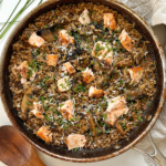 Salmon and mushroom risotto in frying pan with wooden spoon, chives, and napkin surrounding it.