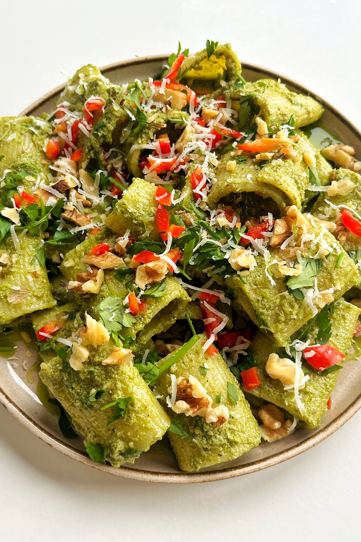 Close up of rigatoni pasta with a zhoug-inspired sauce topped with walnuts, chilli, and parsley on a cream plate and white background.