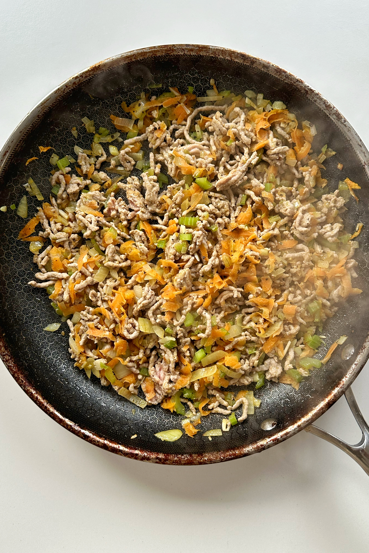 Frying celery, onion, garlic, carrot, and pork mince in a black frying pan on white background.