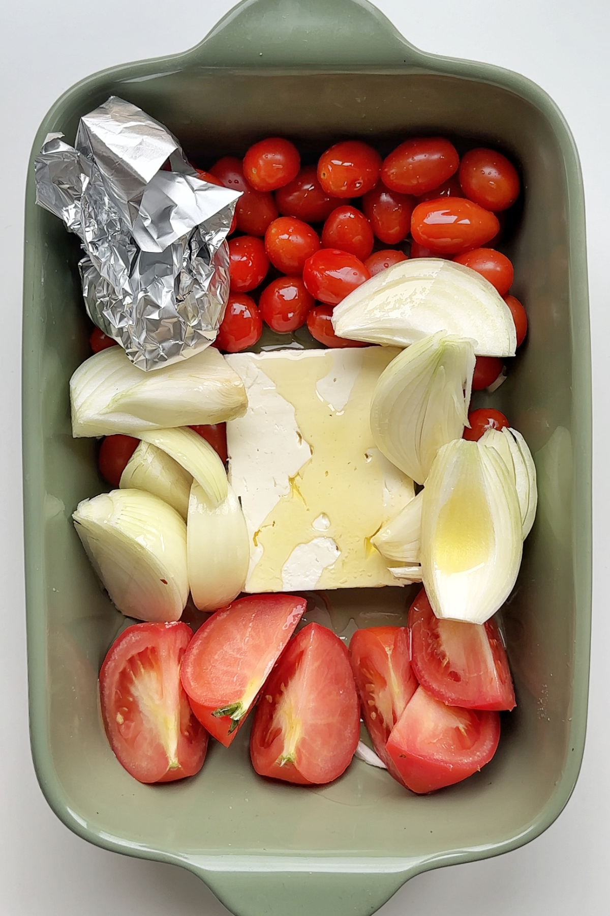 Tomatoes, white onion, feta, and a tinned foiled garlic bulb in a green baking dish.