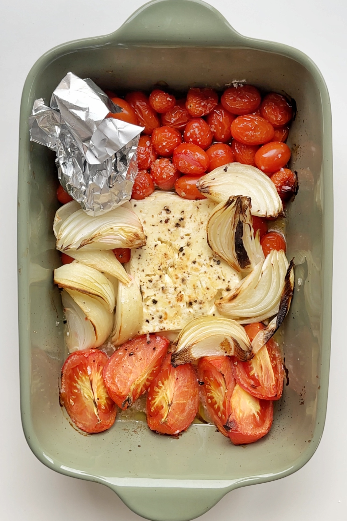Roasted tomatoes, onion, feta, and tinned foiled garlic in a green baking dish.