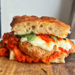 Sandwich with crispy eggplant, mozzarella, and marinara sauce on a chopping board.