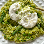 Close up of pea and asparagus orzo with burrata on top on a white plate.