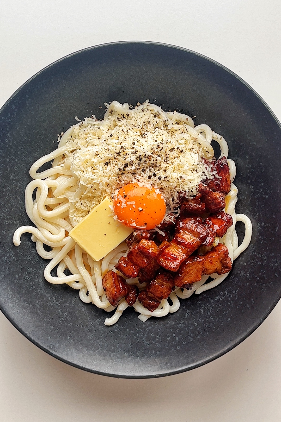 Black bowl with udon noodles, fried pork belly, grated parmesan, knob of butter, and egg yolk.