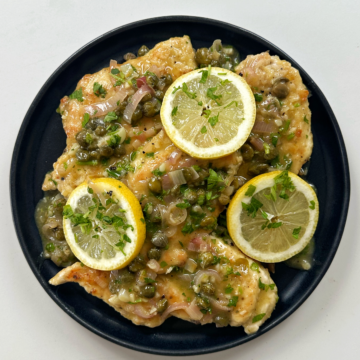 Chicken piccata on a black plate topped with lemon slices.