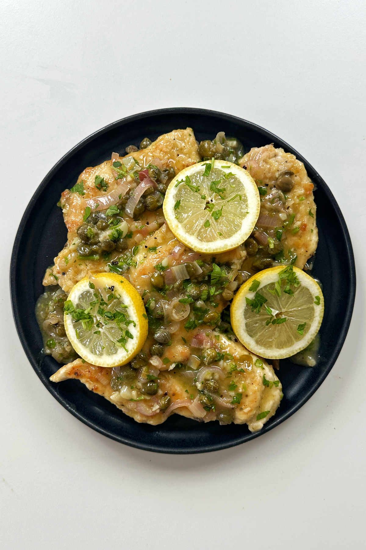 Chicken piccata on a black plate topped with lemon slices. 