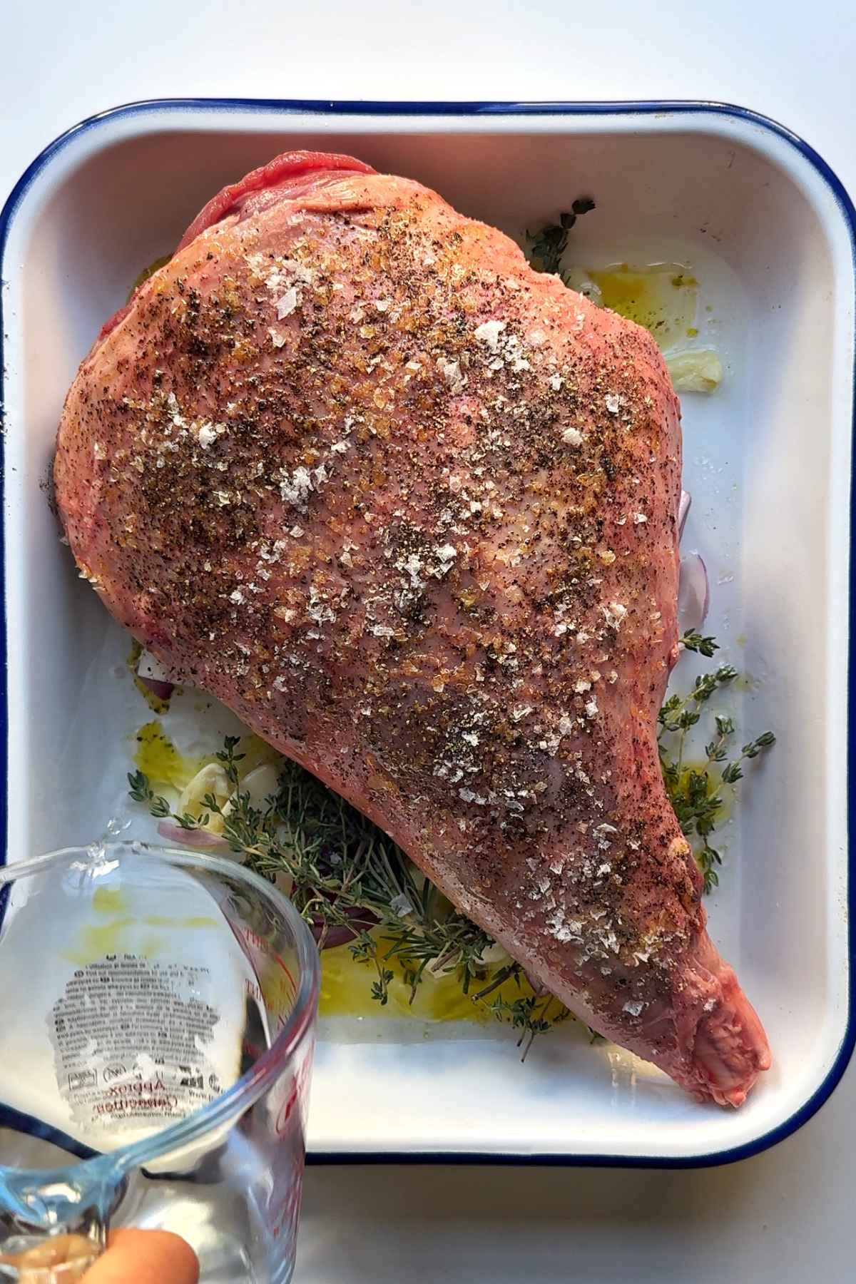 Leg of lamb in tray with thyme, rosemary, onion, and garlic.