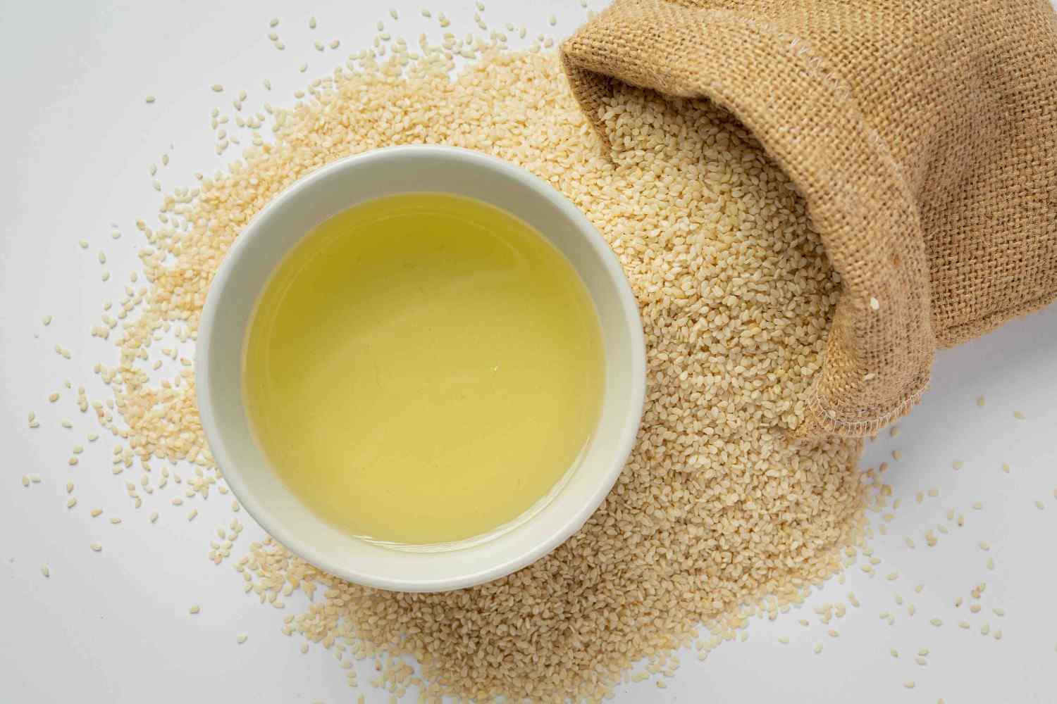 A small bowl of sesame oil next to a back of spilled sesame seeds