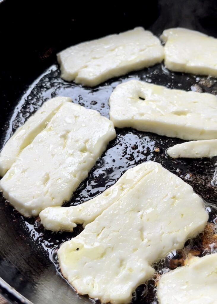 Slices of halloumi cheese being pan-fried in a skillet until they are crispy and golden brown on the outside, serving as a salty and addictive ramen topping that mimics the function of traditional Agedashi Tofu.