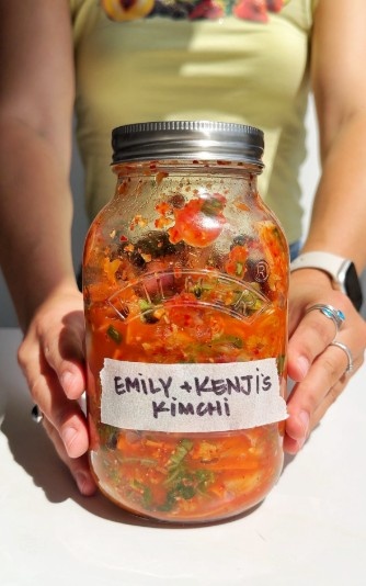 A large glass jar filled with vibrant, red homemade kimchi and labeled with a handwritten "Emily + Kenji's Kimchi" sticker, held by hands against a bright background to be used as a fermented ramen topping.