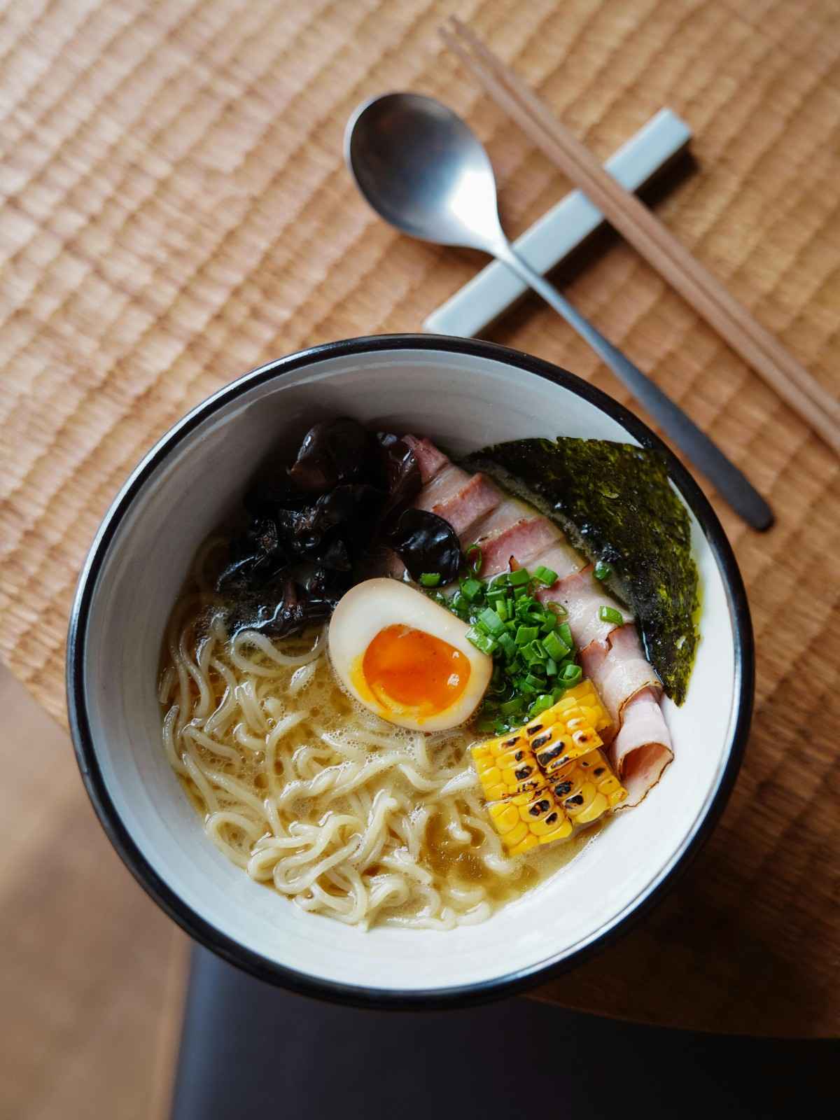 A top-down view of a ramen bowl featuring a piece of charred sweet corn alongside wood ear mushrooms, pork chashu, and a jammy soft-boiled egg.