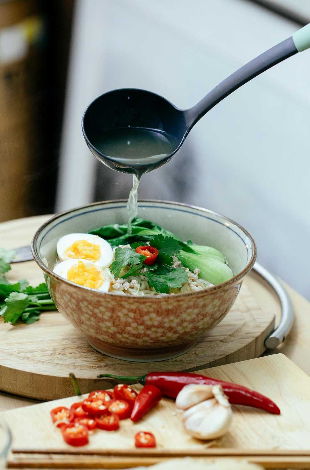 Clear broth being poured from a ladle into a floral ceramic bowl of ramen topped with vibrant green baby bok choy, halved soft-boiled eggs, and fresh cilantro, demonstrating the "one-pot" steaming method for leafy greens.