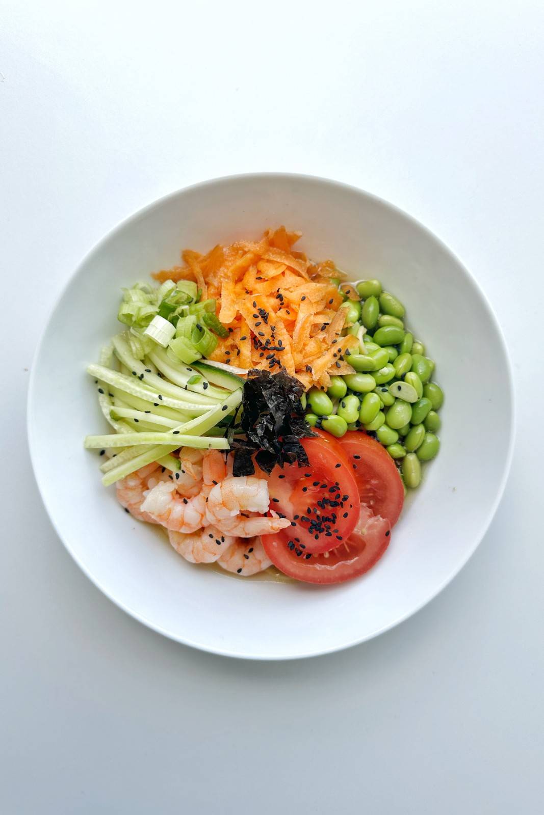 A top-down view of a white bowl of chilled noodles topped with pink prawns, edamame beans, sweet sliced tomatoes, julienned cucumber, shredded carrots, and spring onions, finished with black sesame seeds. This illustrates the "Cold Garnishments" hack for a refreshing 10-minute summer ramen.
