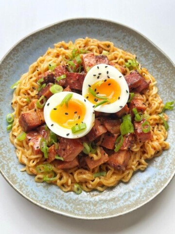 A bowl of instant ramen noodles topped with caramelized cubes of fried Spam, a halved soft-boiled egg, and sliced green onions, showcasing the viral Spam hack.