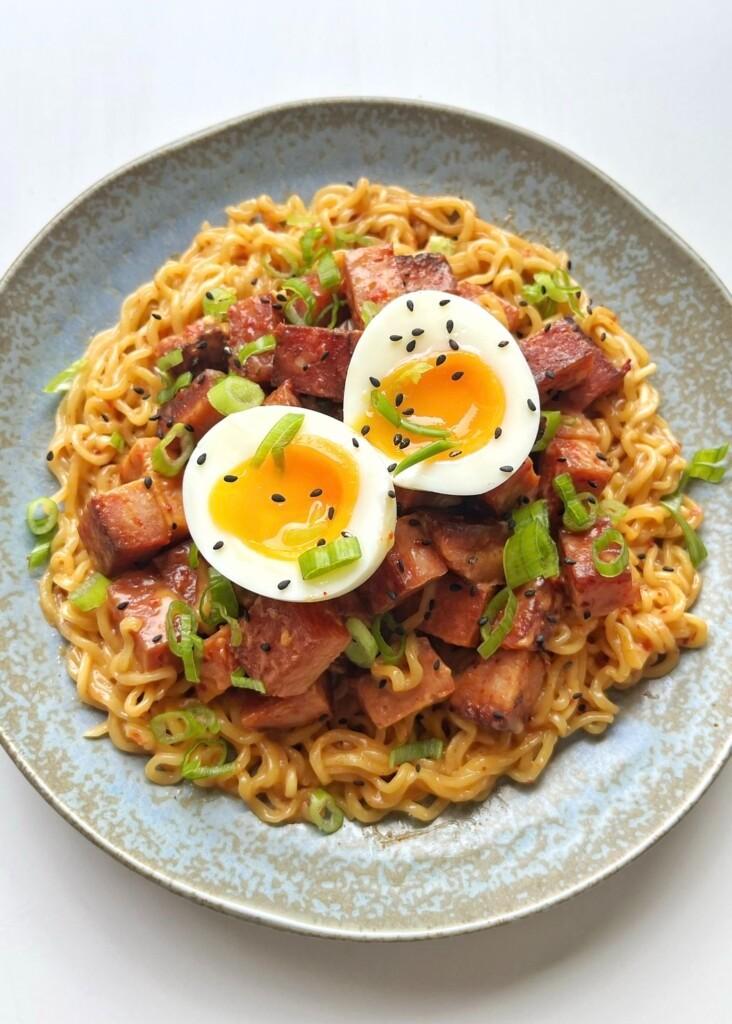 A bowl of instant ramen noodles topped with caramelized cubes of fried Spam, a halved soft-boiled egg, and sliced green onions, showcasing the viral Spam hack.