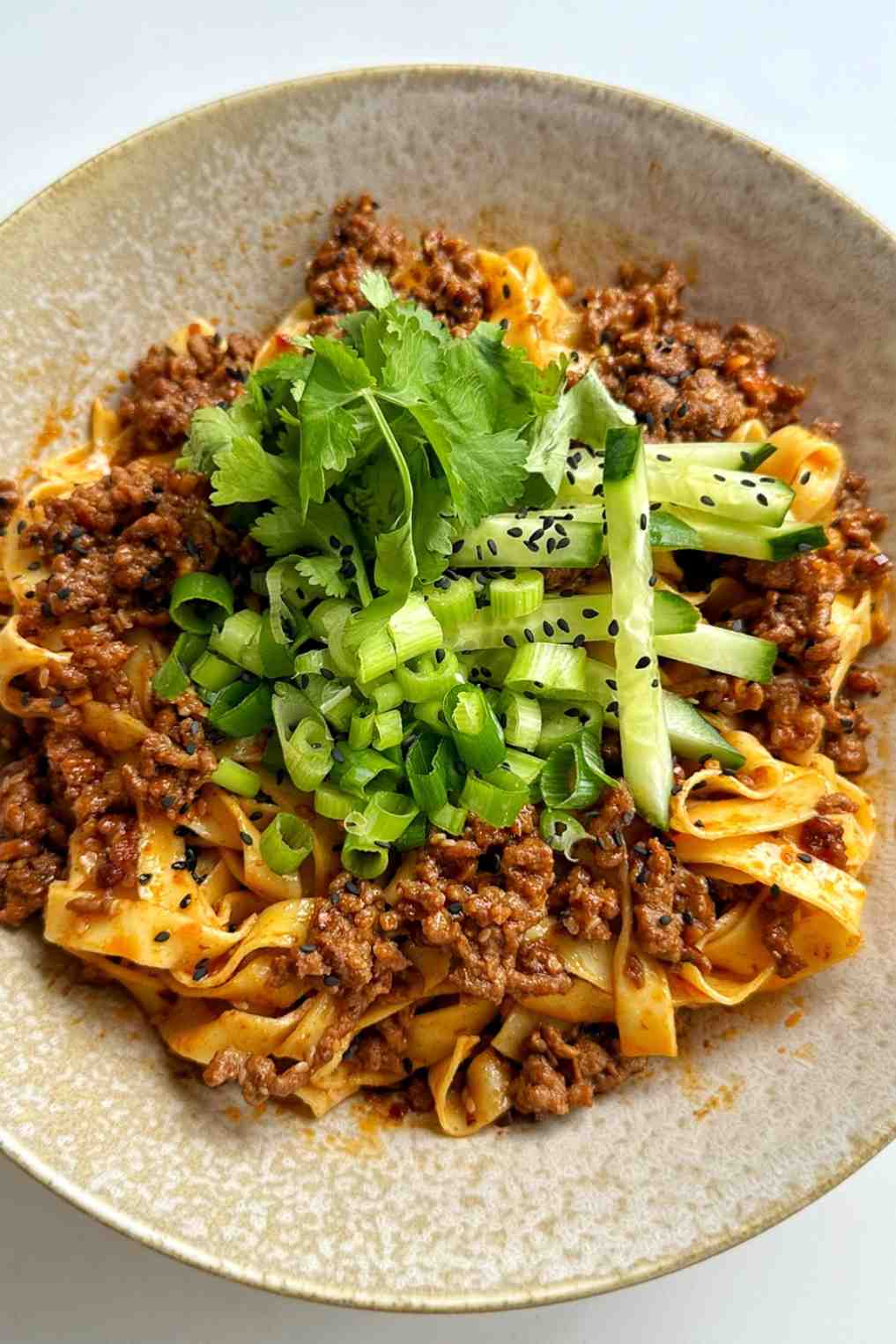 A bowl of flat noodles tossed with crispy ground beef, with sliced green onions, fresh cilantro, julienned cucumber, and black sesame seeds, as described in the crispy ground beef section.