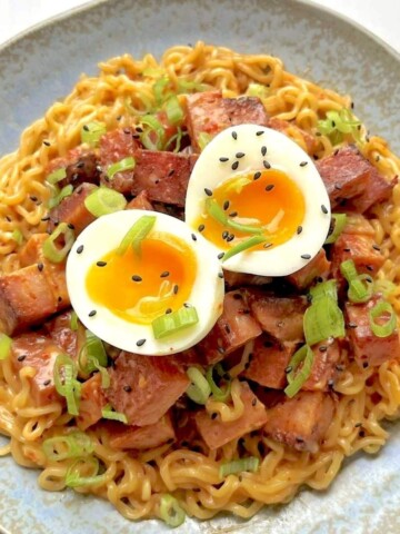 A bowl of instant ramen noodles topped with caramelized cubes of fried Spam, a halved soft-boiled egg, and sliced green onions, showcasing the viral Spam hack.