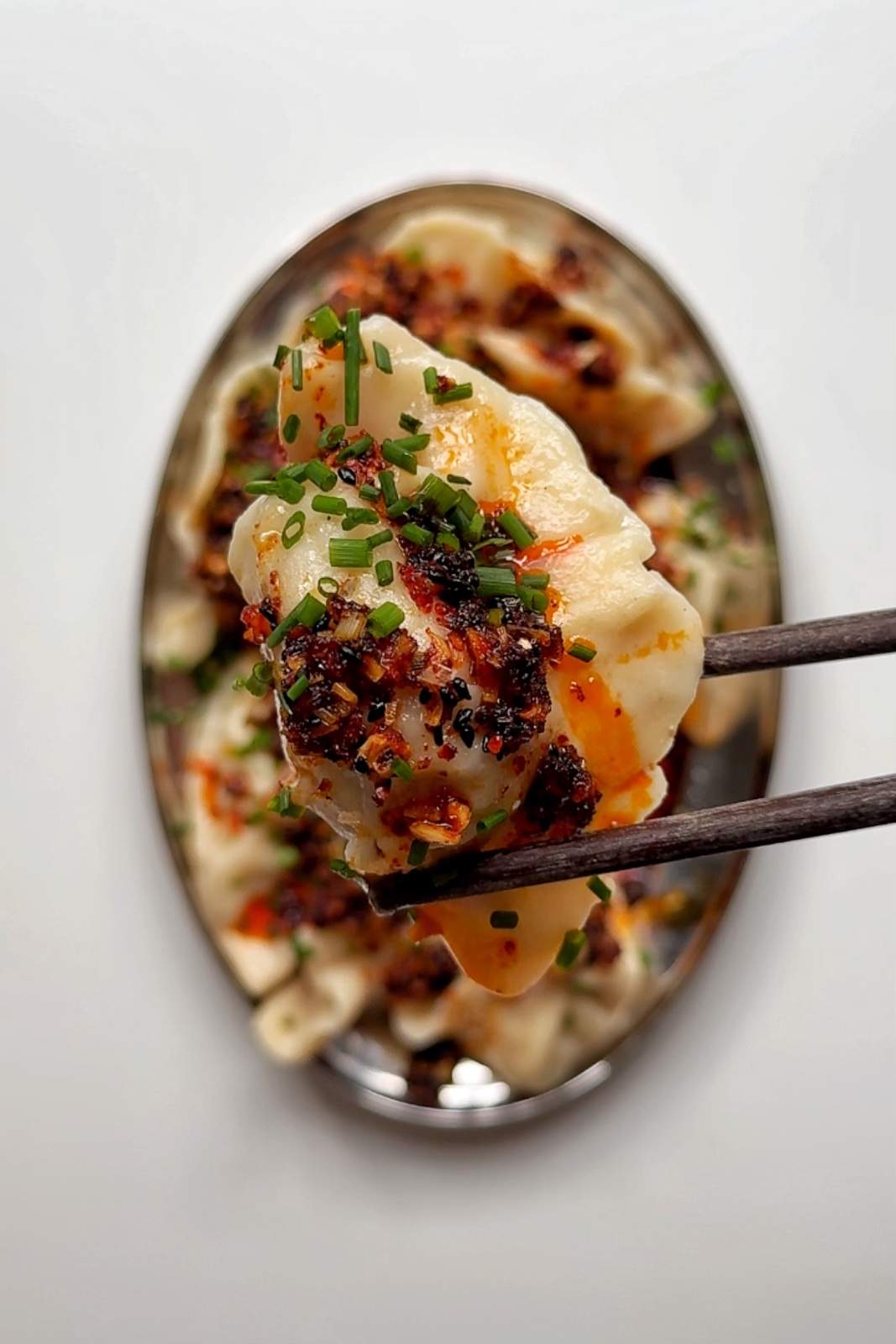 A close-up of a gyoza dumpling held with chopsticks, topped with spicy chili crunch and fresh chives.