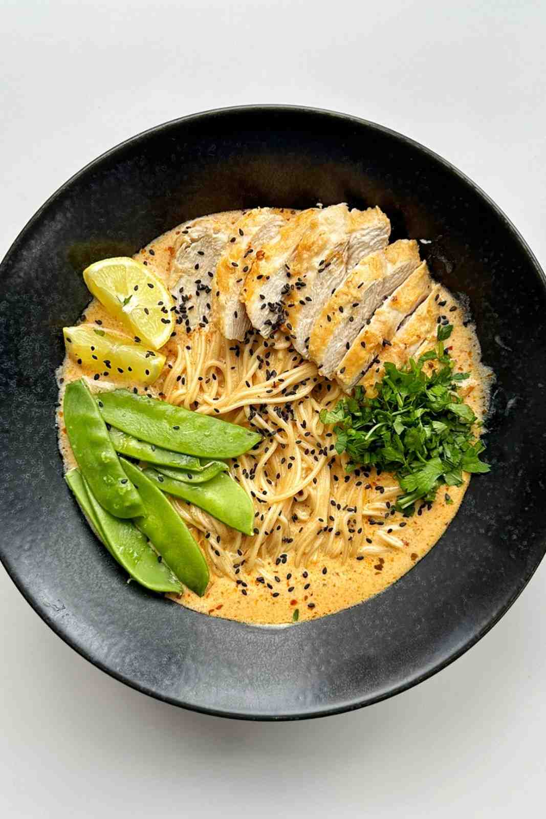 Sliced rotisserie chicken served in a bowl of creamy lemon chicken ramen with snow peas, fresh herbs, and lemon wedges.