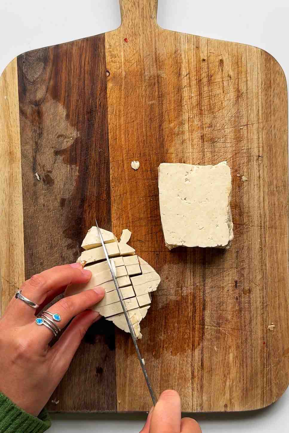 Hands dicing a block of firm tofu on a wooden cutting board, demonstrating the "tofu sponge" preparation method.