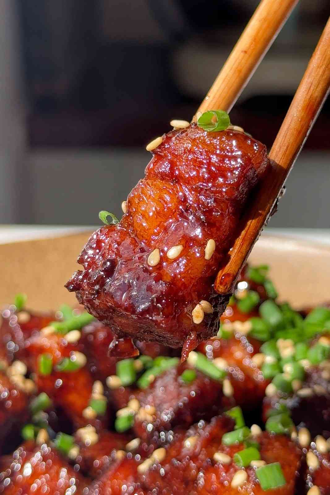 A close-up of a piece of tender, glazed Asian braised pork belly held with chopsticks, topped with sesame seeds and spring onions, serving as a premium ramen topping.