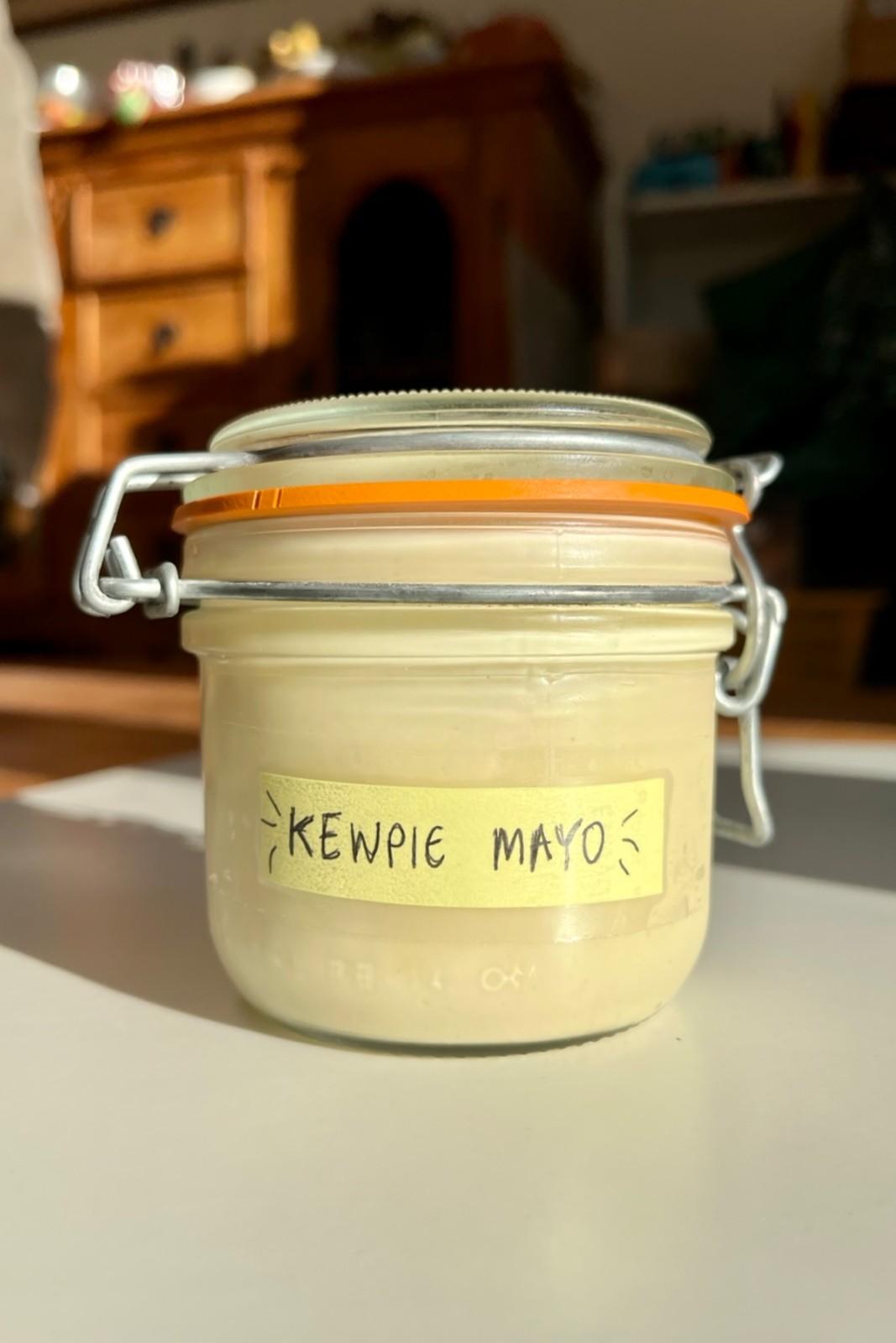 A close-up of a glass jar filled with homemade Kewpie mayonnaise, labeled with a handwritten sticker, used for the "Kewpie Swirl" ramen broth hack.